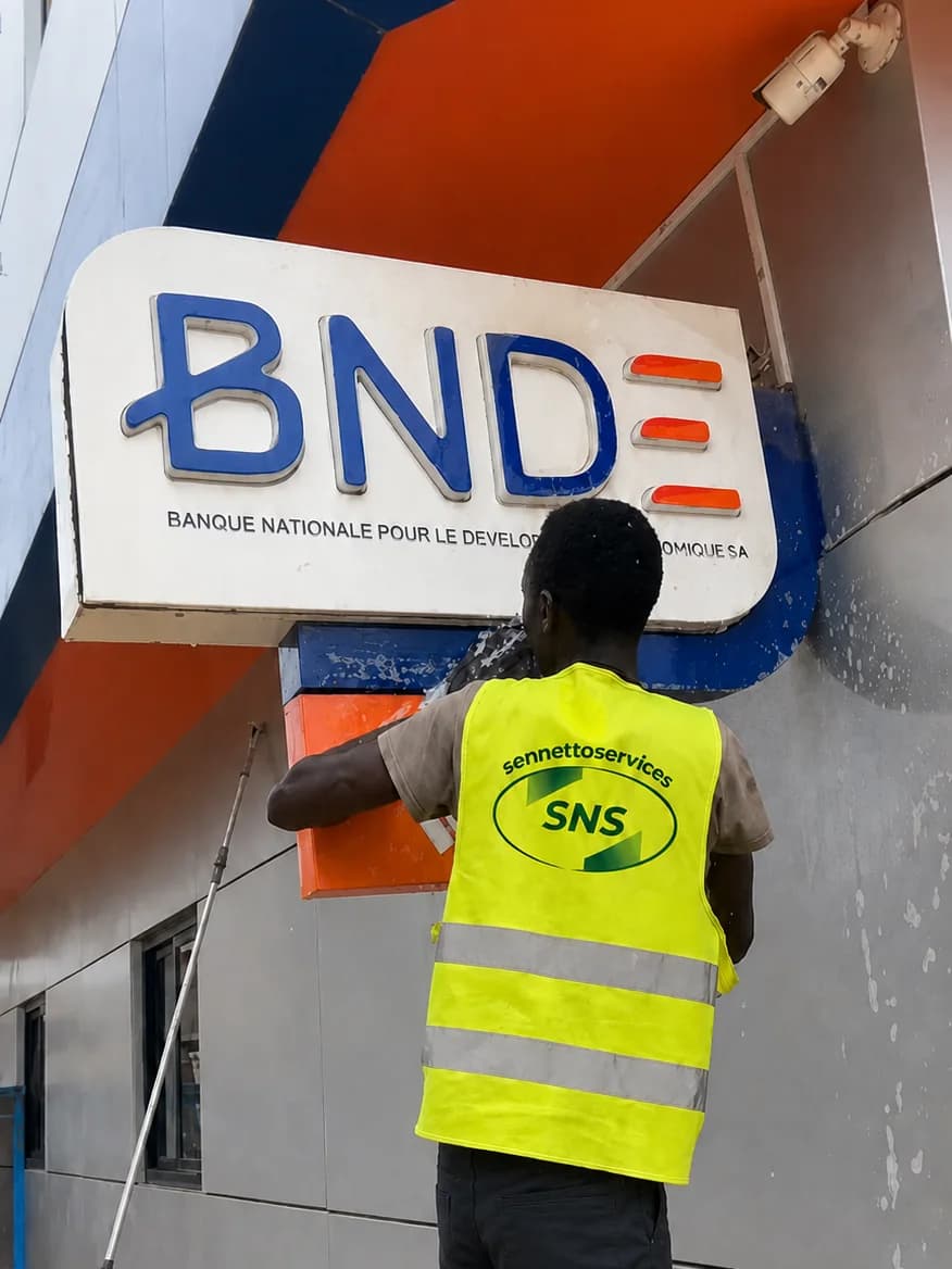 Agent SenNettoServices en gilet SNS nettoyant l'enseigne de la BNDE à Dakar